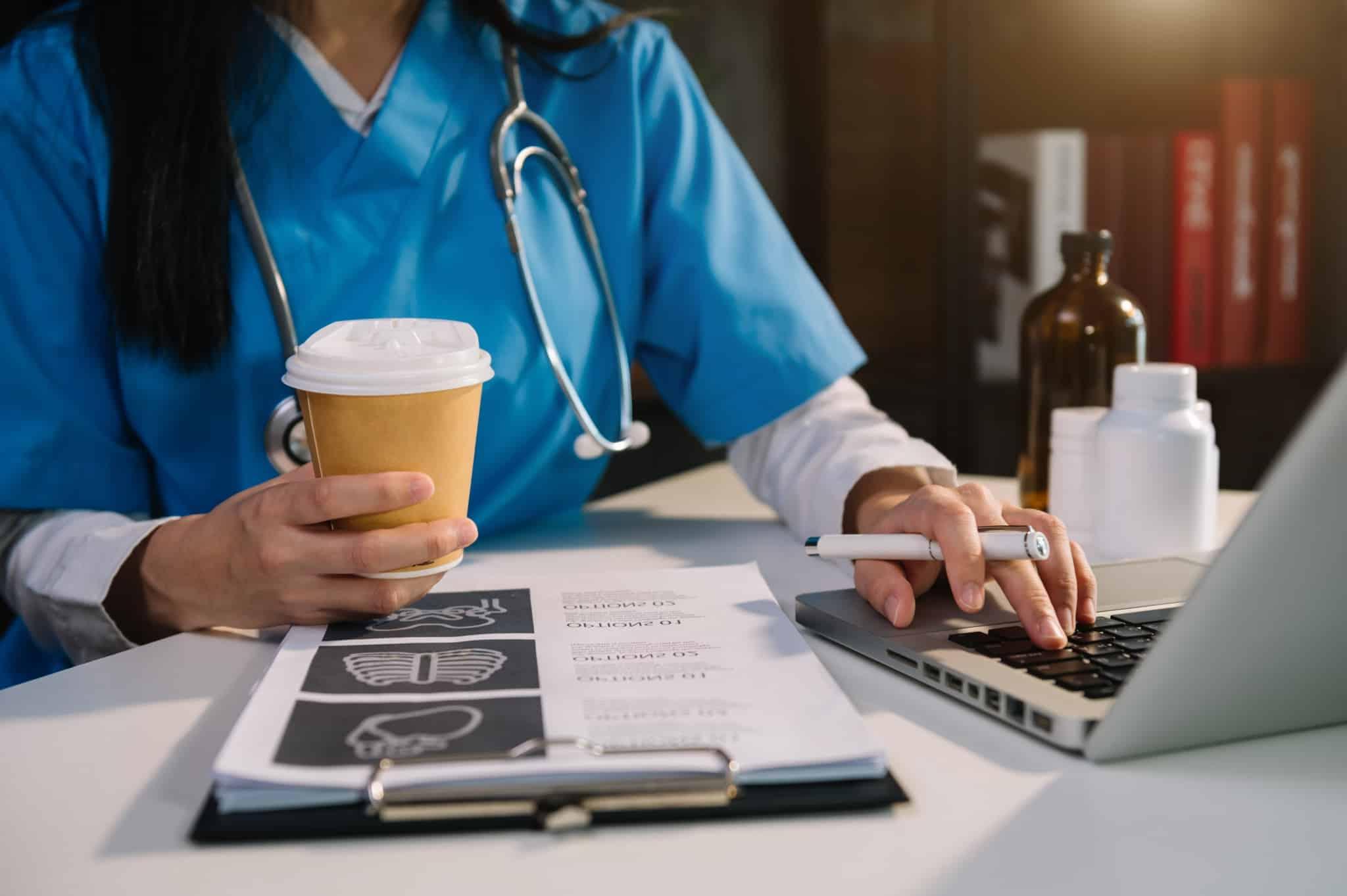 Medical professional holding a cup of coffee while typing on a laptop